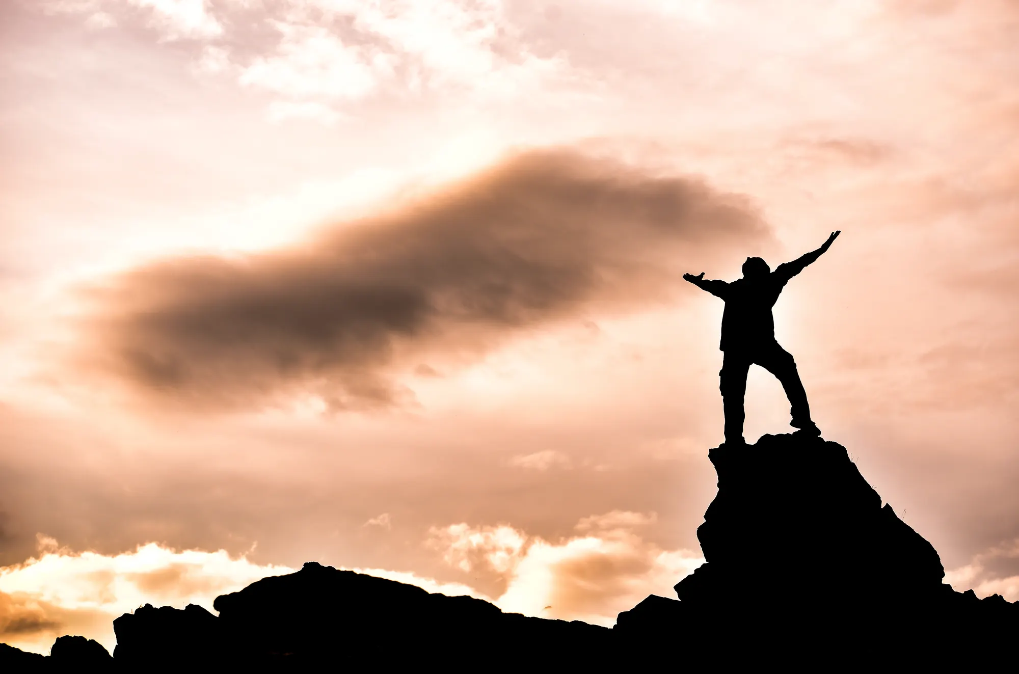 Silhouette of a person on a mountain peak, arms raised, against a dramatic sunset sky