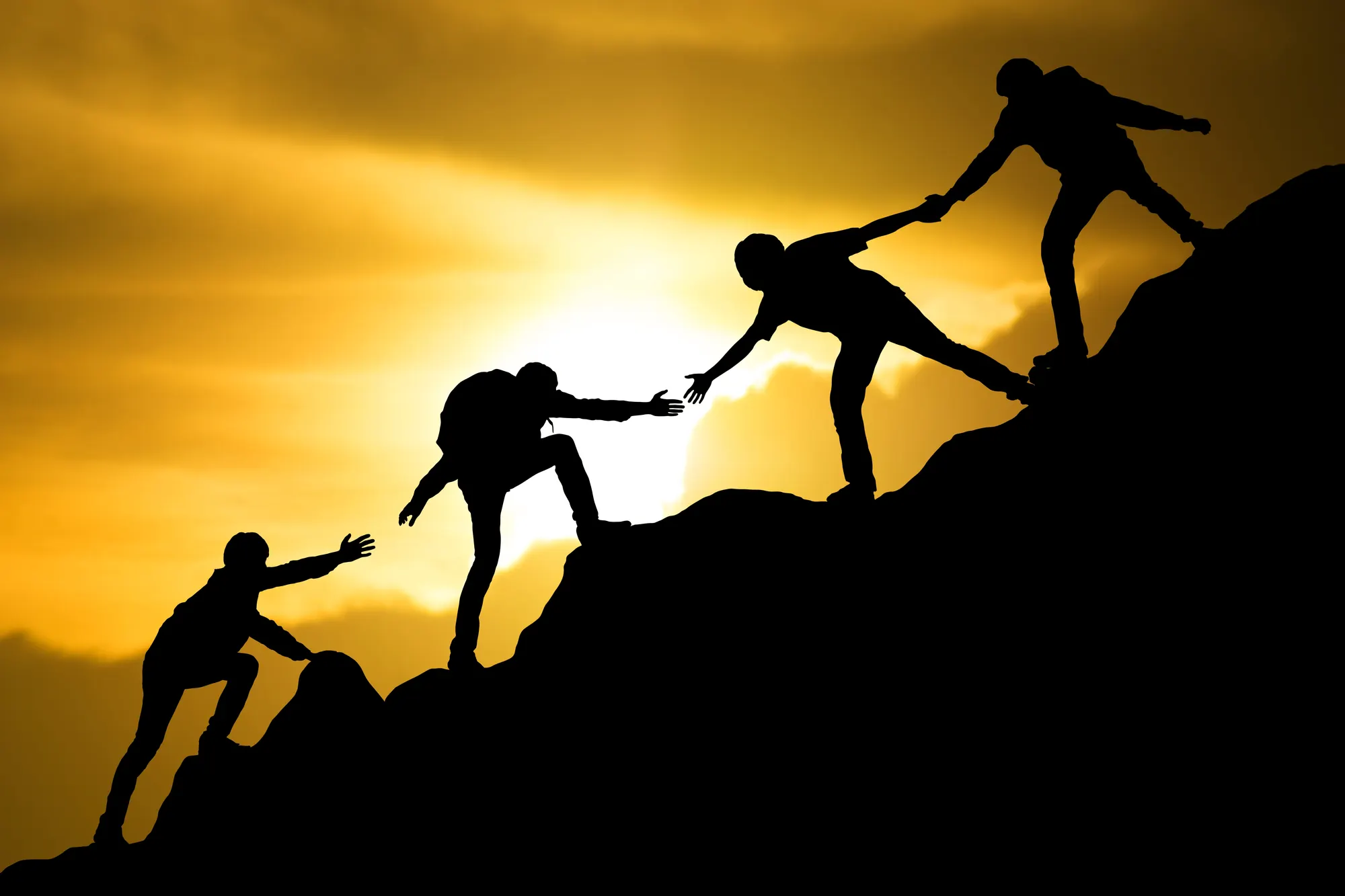 Four hikers helping each other climb a mountain ridge at sunset